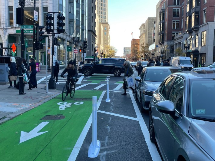 A person rides a bike on a freshly-painted green bike lane next to some flexible-post bollards on a busy multi-lane city street lined with tall buildings