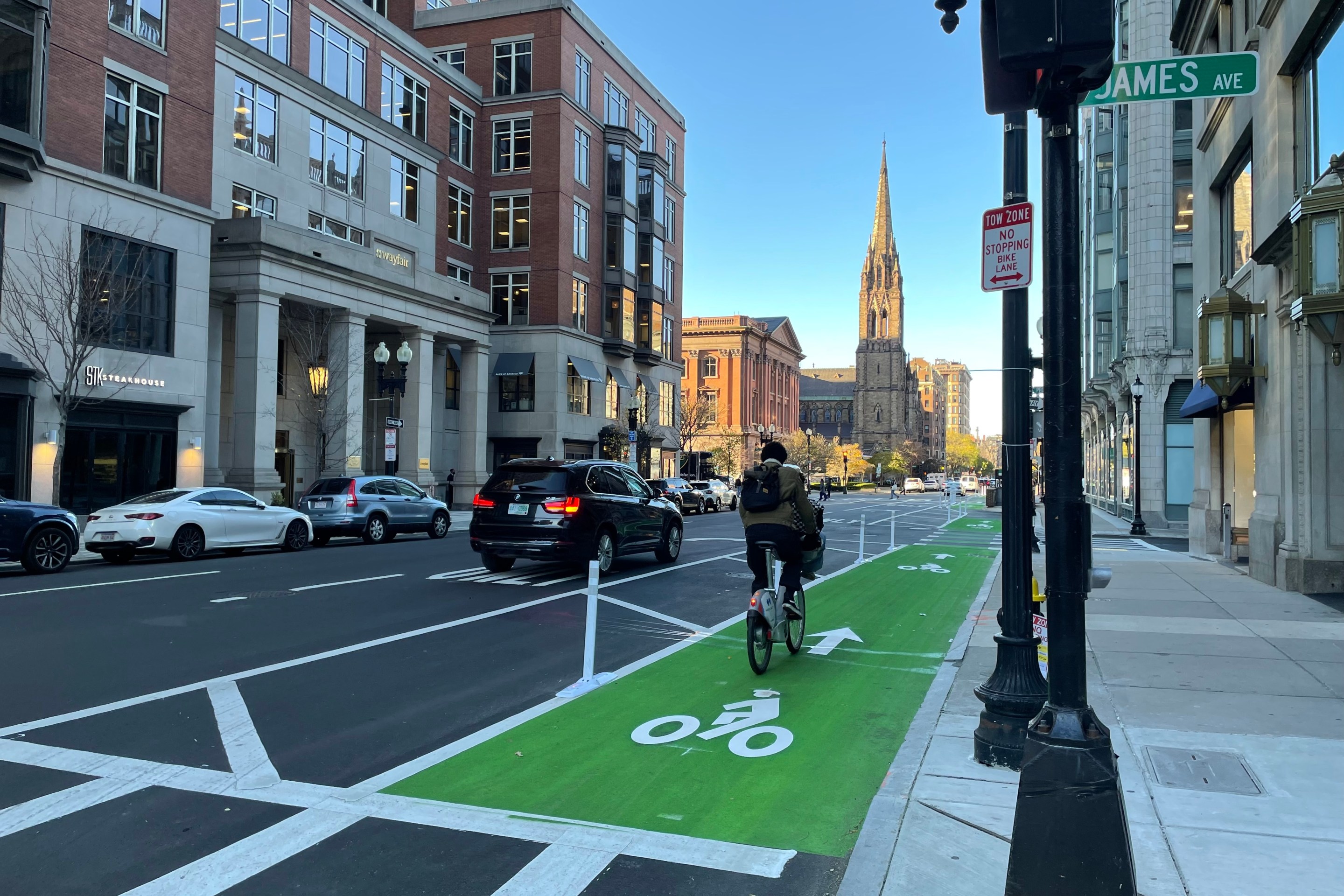 A person rides a bike on a freshly-painted green bike lane next to some flexible-post bollards on a multi-lane city street lined with tall buildings