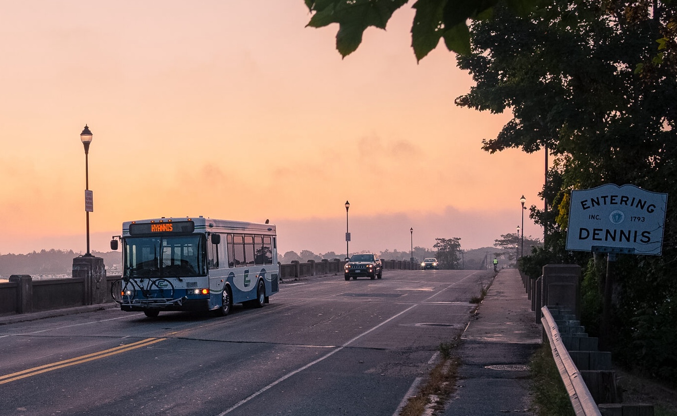 A bus with "HYANNIS" on its destination sign crosses a bridge under a pink sky at sunrise next to a sign that says "ENTERING DENNIS"