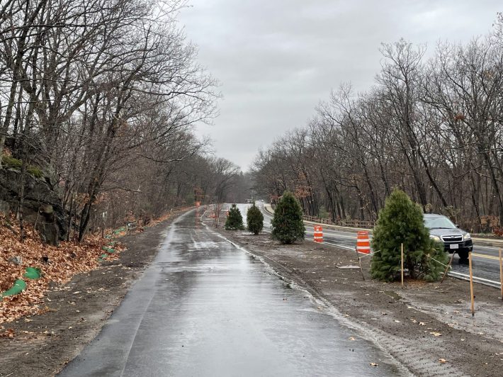 A bike path runs parallel next two a two-lane roadway running through a forest. Between the bike path and the roadway are some orange construction barrels and some newly-planted trees in a 10-foot-wide strip.