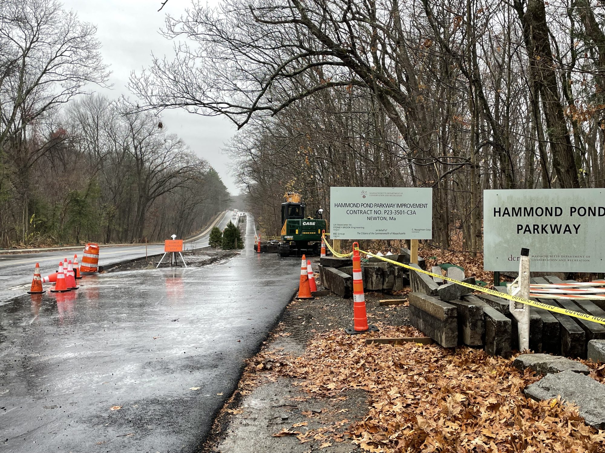 Signs alongside a roadway read "Hammond Pond Parkway – Massachusetts Dept. of Conservation and Recreation" and "Hammond Pond Parkway Improvement Contract No. P23-3501-C3A Newton MA" next to some orange construction cones and freshly-laid asphalt and stacks of granite curbing.