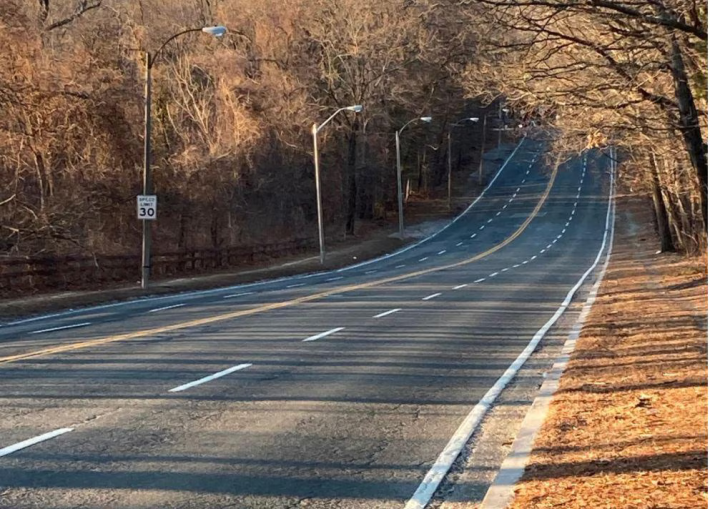 A straight four-lane highway with no sidewalks runs through a forest.