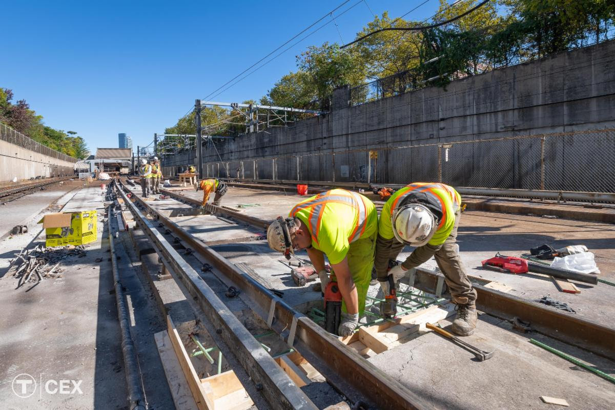 Workers in dayglo vests work on train tracks on a concrete platform under a bright blue sky.