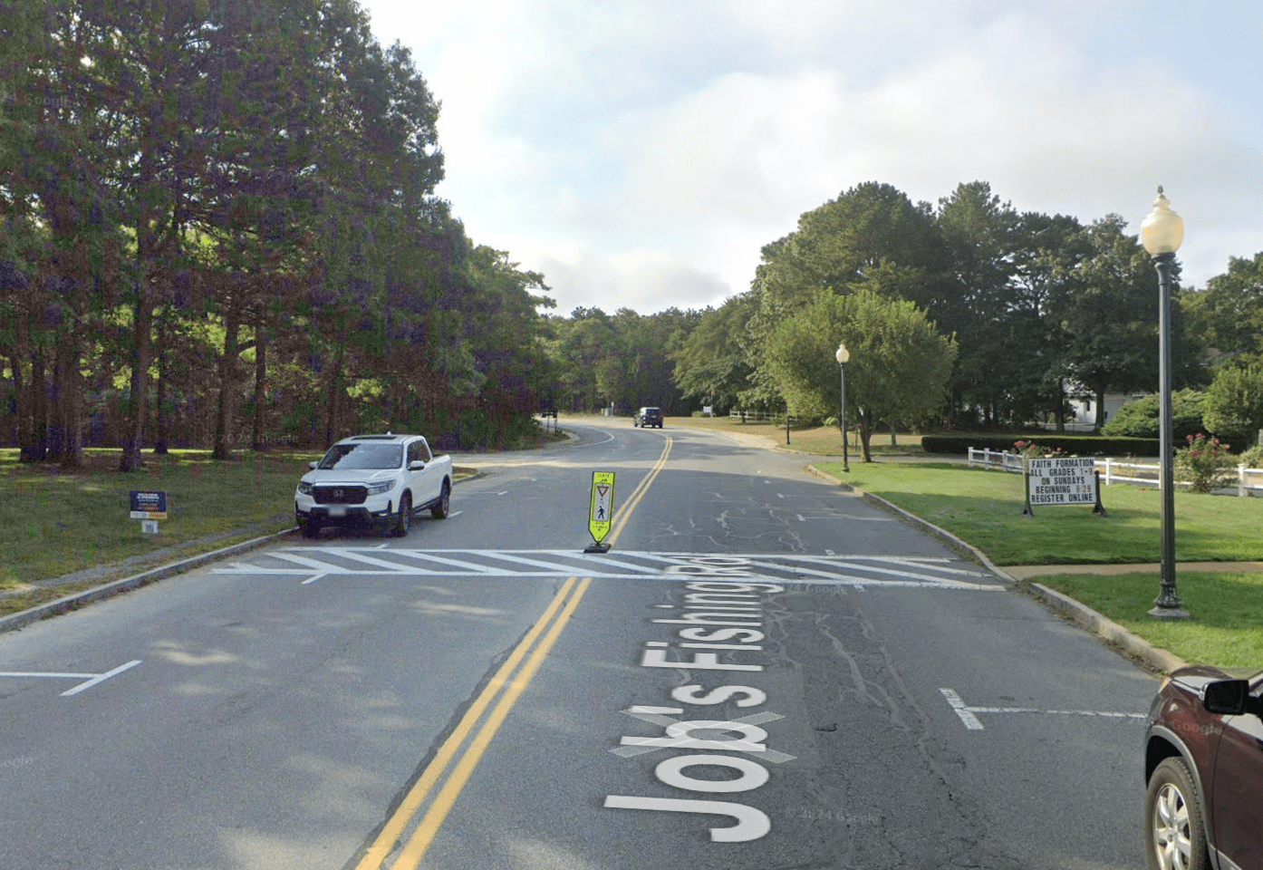 A Google Street View image of a two-lane street with on-street parking on either side, and a well-marked crosswalk in the middle distance. There are no sidewalks on the edges of the street.