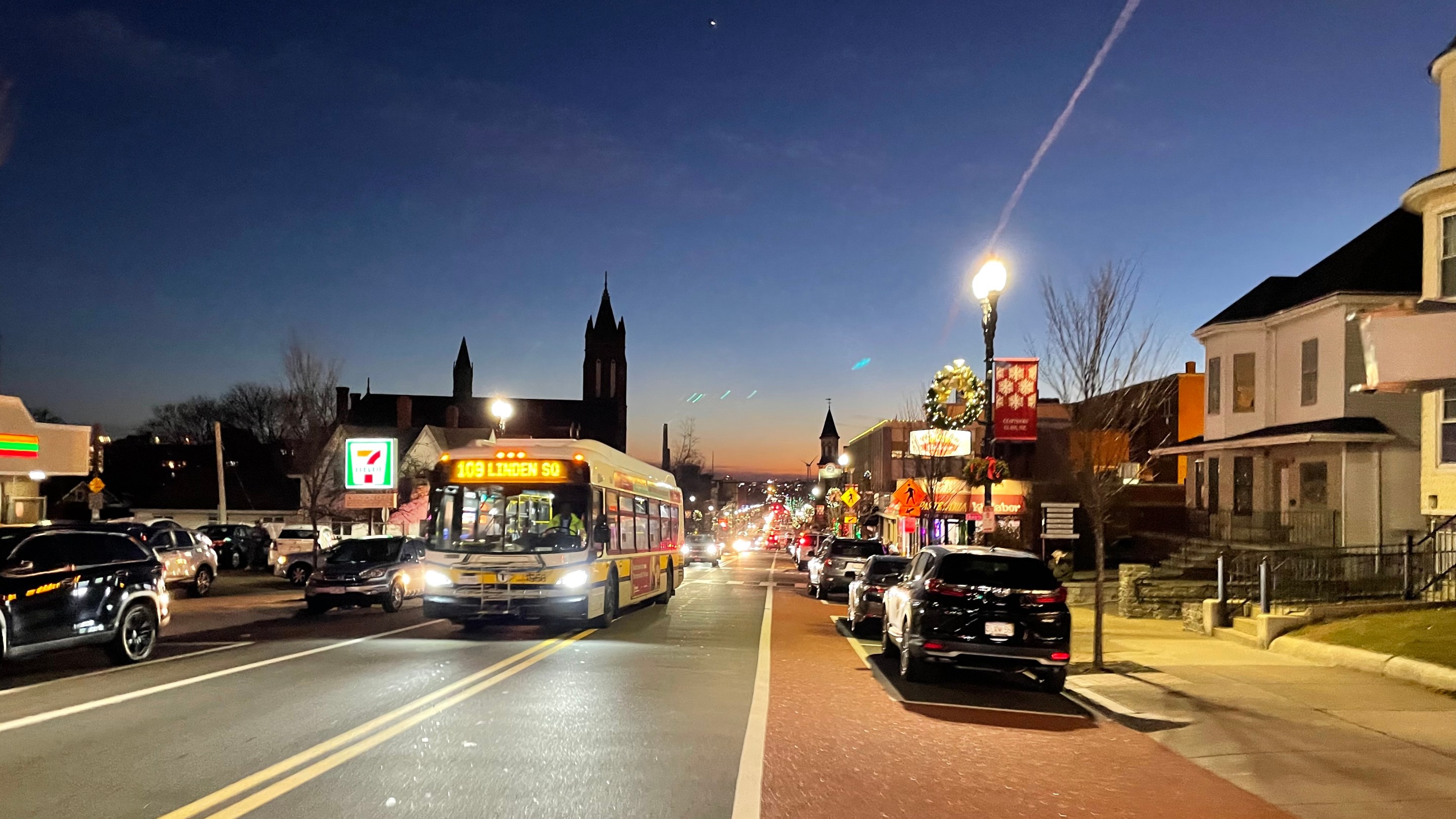 A bus drives up a long, straight city street lined with parked cars and 2-3 story buildignsat dusk.