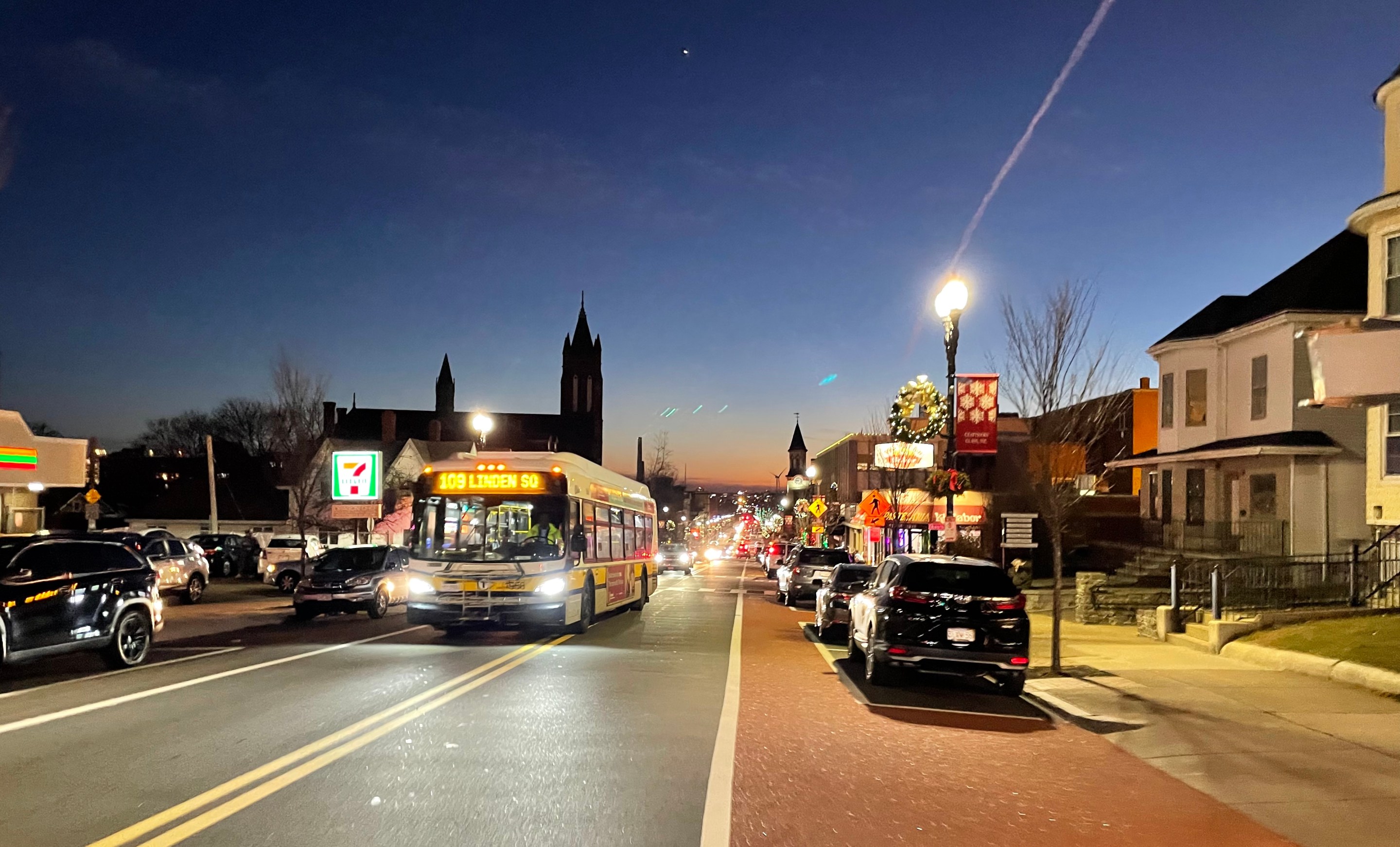 A bus drives up a long, straight city street lined with parked cars and 2-3 story buildignsat dusk.