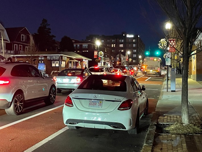 A white Mercedes sedan blocks a bus lane next to a long line of stopped cars. In the middle distance is an MBTA bus stopped at a curbside bus stop.