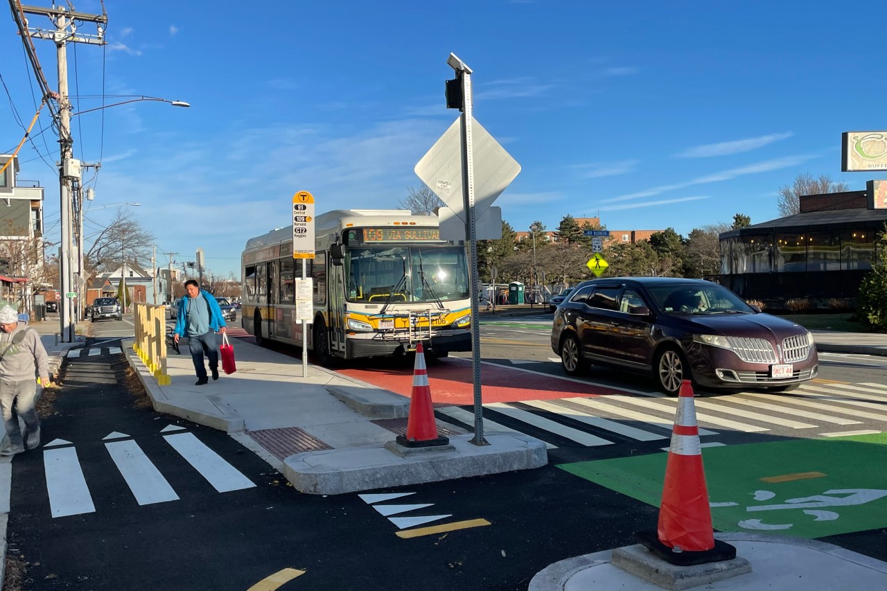 A man walks away from a bus stop where an MBTA bus marked "109 Via Sullivan" is waiting. In the foreground is a crosswalk.