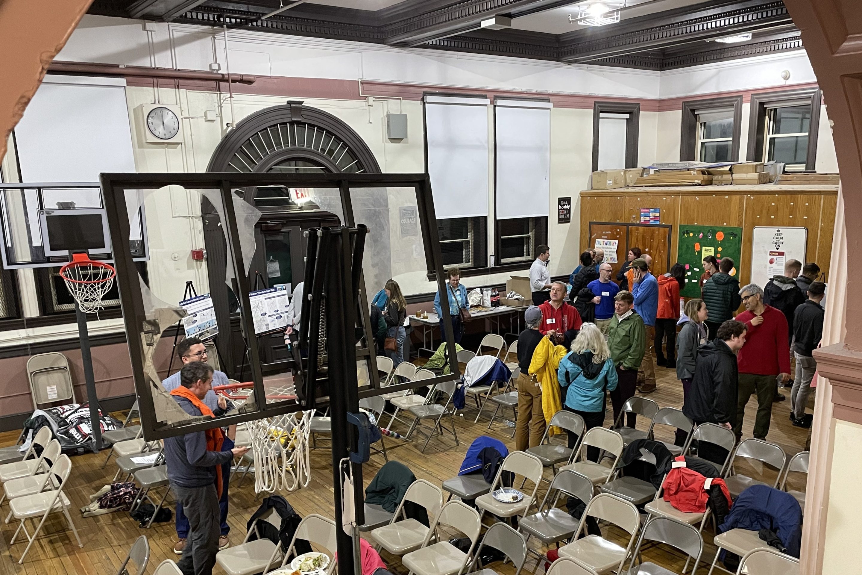 A few dozen people in rain coats chat in small groups inside a school gymnasium filled with folding chairs.