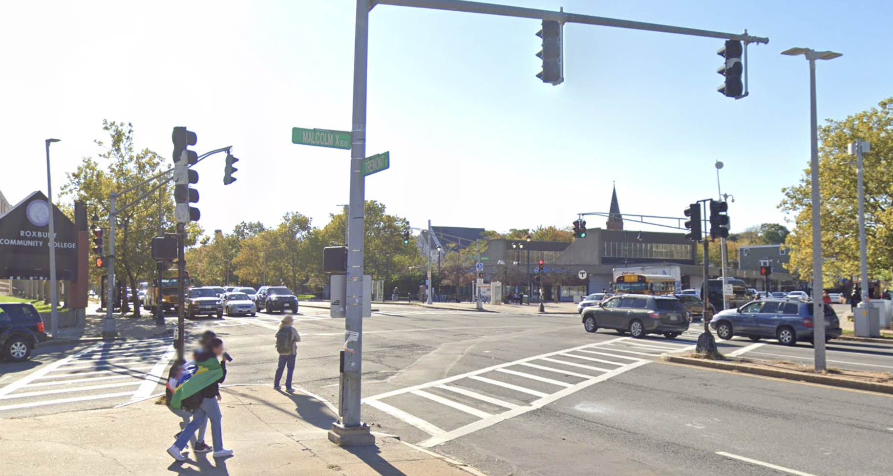 Pedestrians wait to cross a wide 6-lane highway at a traffic light. In the distance on the far side of the intersection is a T station.