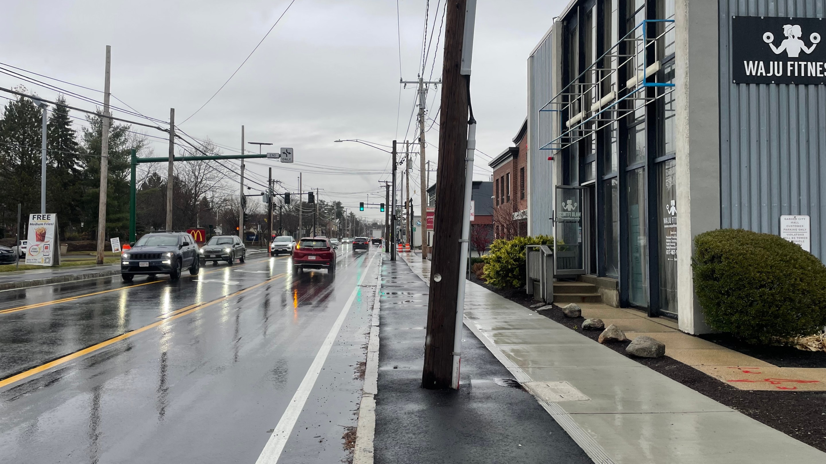 A freshly-paved city street lined with commercial buildings against the sidewalk. In the center of the image, a freshly-paved sidewalk-level bike lane is blocked by a utility pole.