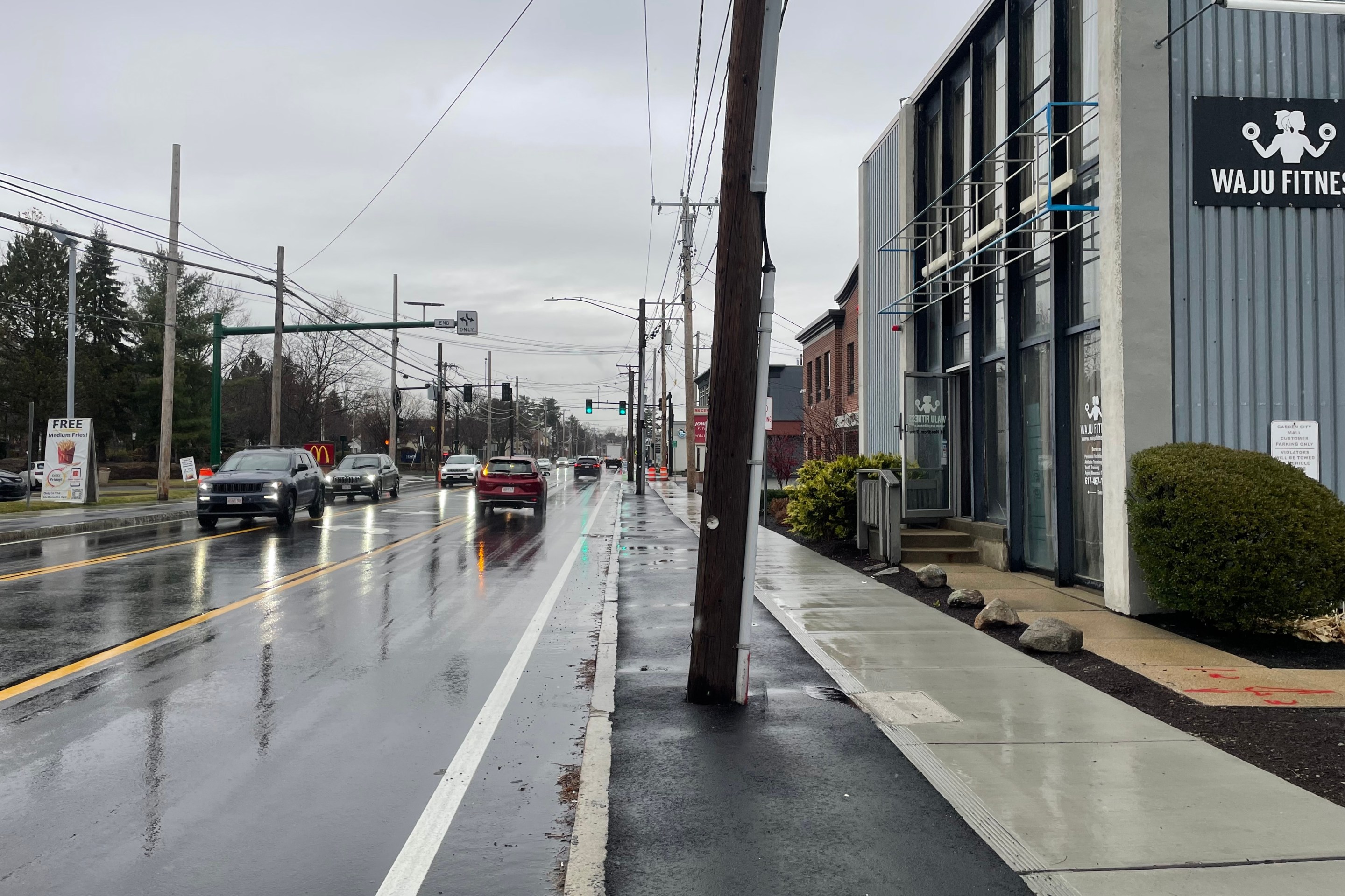 A freshly-paved city street lined with commercial buildings against the sidewalk. In the center of the image, a freshly-paved sidewalk-level bike lane is blocked by a utility pole.