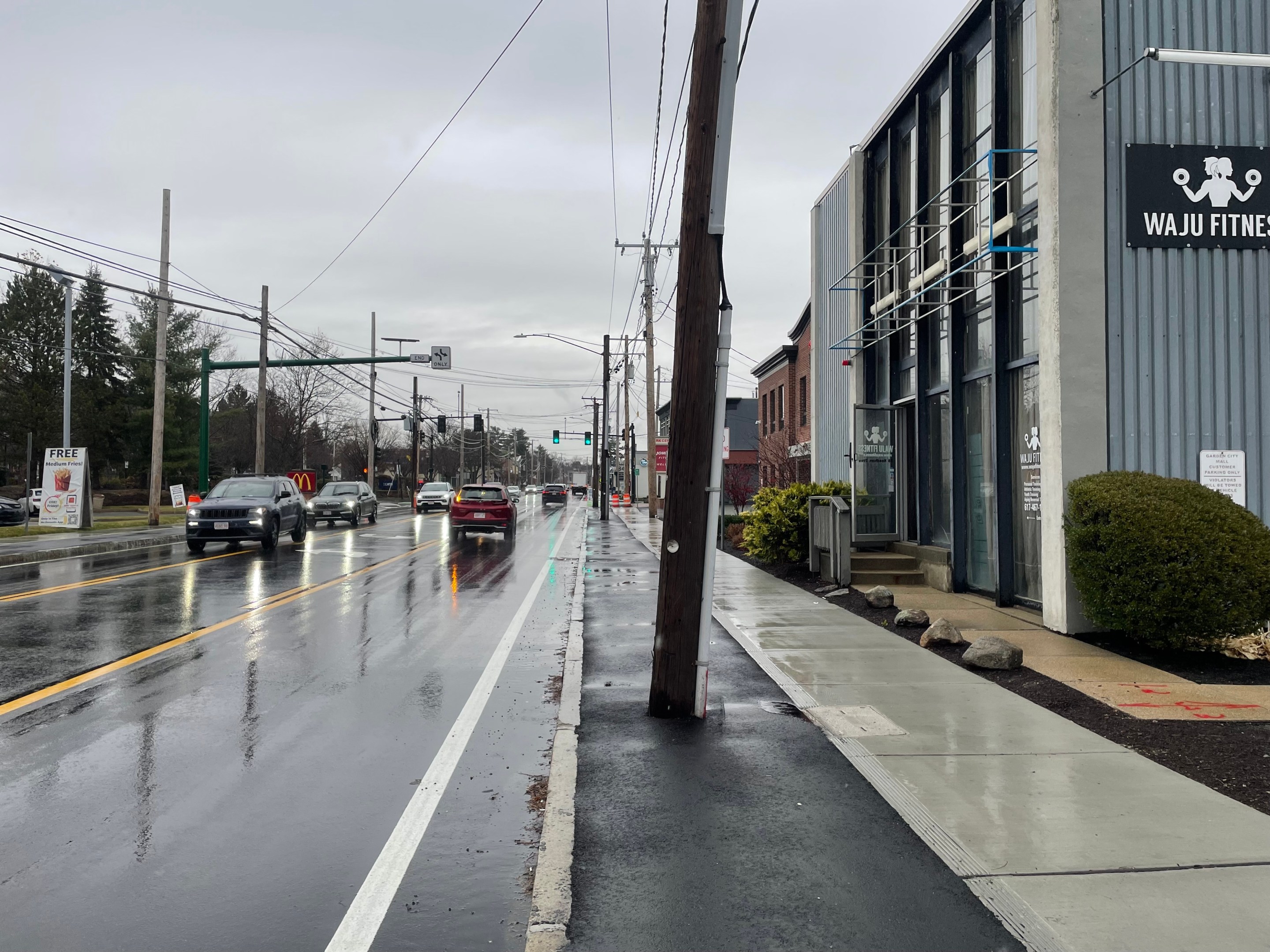 A freshly-paved city street lined with commercial buildings against the sidewalk. In the center of the image, a freshly-paved sidewalk-level bike lane is blocked by a utility pole.