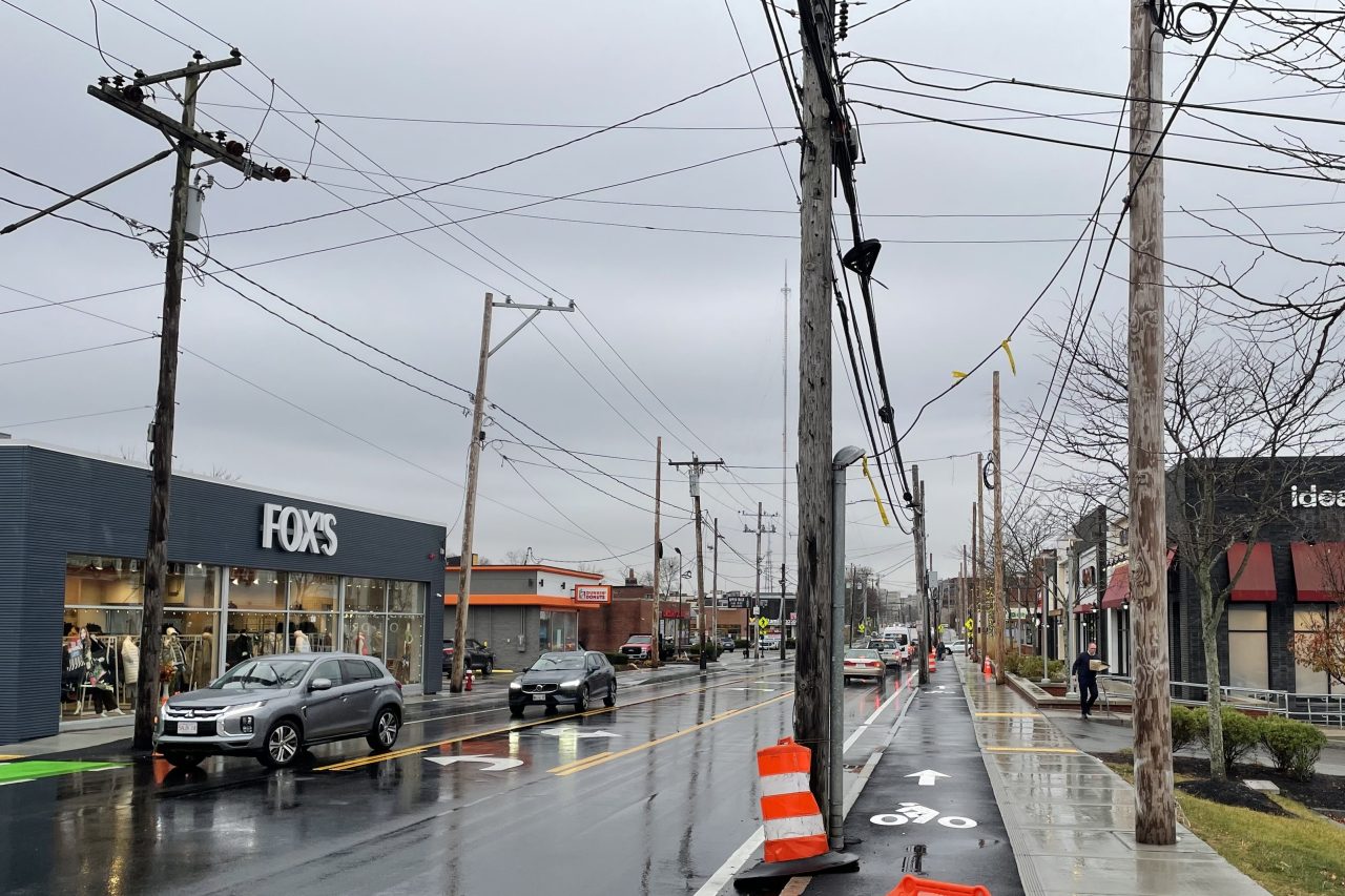 Dozens of wires suspended over a street lined with small businesses and orange construction barrels. The photo shows four rows of utility poles along the street. In the center, pairs of poles are located on the outside of the sidewalk and in the curb between the street's bike lane and roadway; on the left edge of the photo on the other side of the street are more poles.