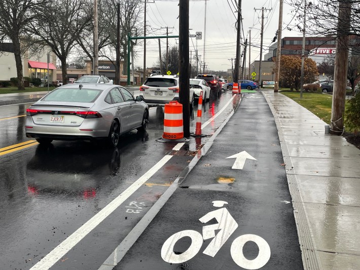 A freshly-paved city street lined with commercial buildings. In the center of the image there is a freshly-paved sidewalk-level bike lane. In the middle of the photo two utility poles block the edge of the motor vehicle lane. There are orange construction barrels at the base of both poles.