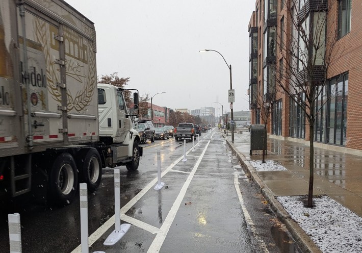 A truck passes a bike lane separated from the traffic lane by a row of plastic flexible-post bollards. The pavement is wet with falling snow, which is beginning to accumulate in the planting areas of the adjacent sidewalk.