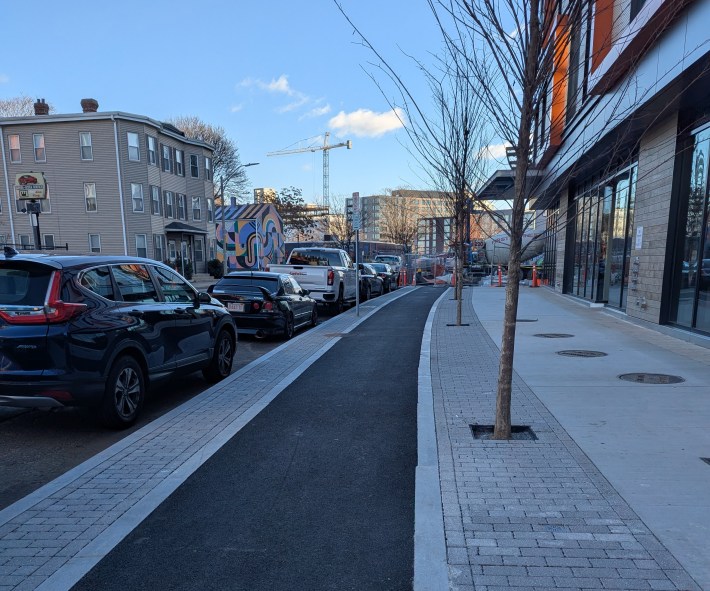 A sidewalk-level bike lane runs alongside a new sidewalk next to a new office building with a row of parked cars on the street to the left. A crane rises on the horizon in the distance.