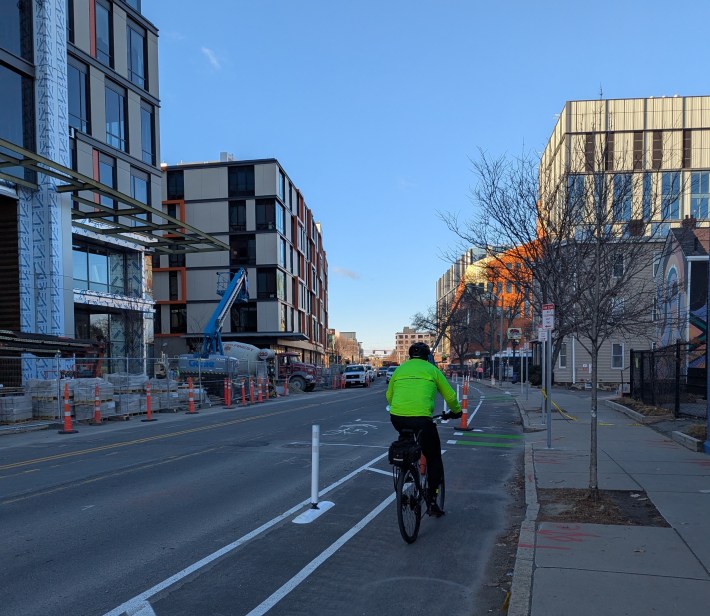 A rider wearing a dayglo yellow jacket rides in a bike lane next to a construction site for a large multi-story lab office building.