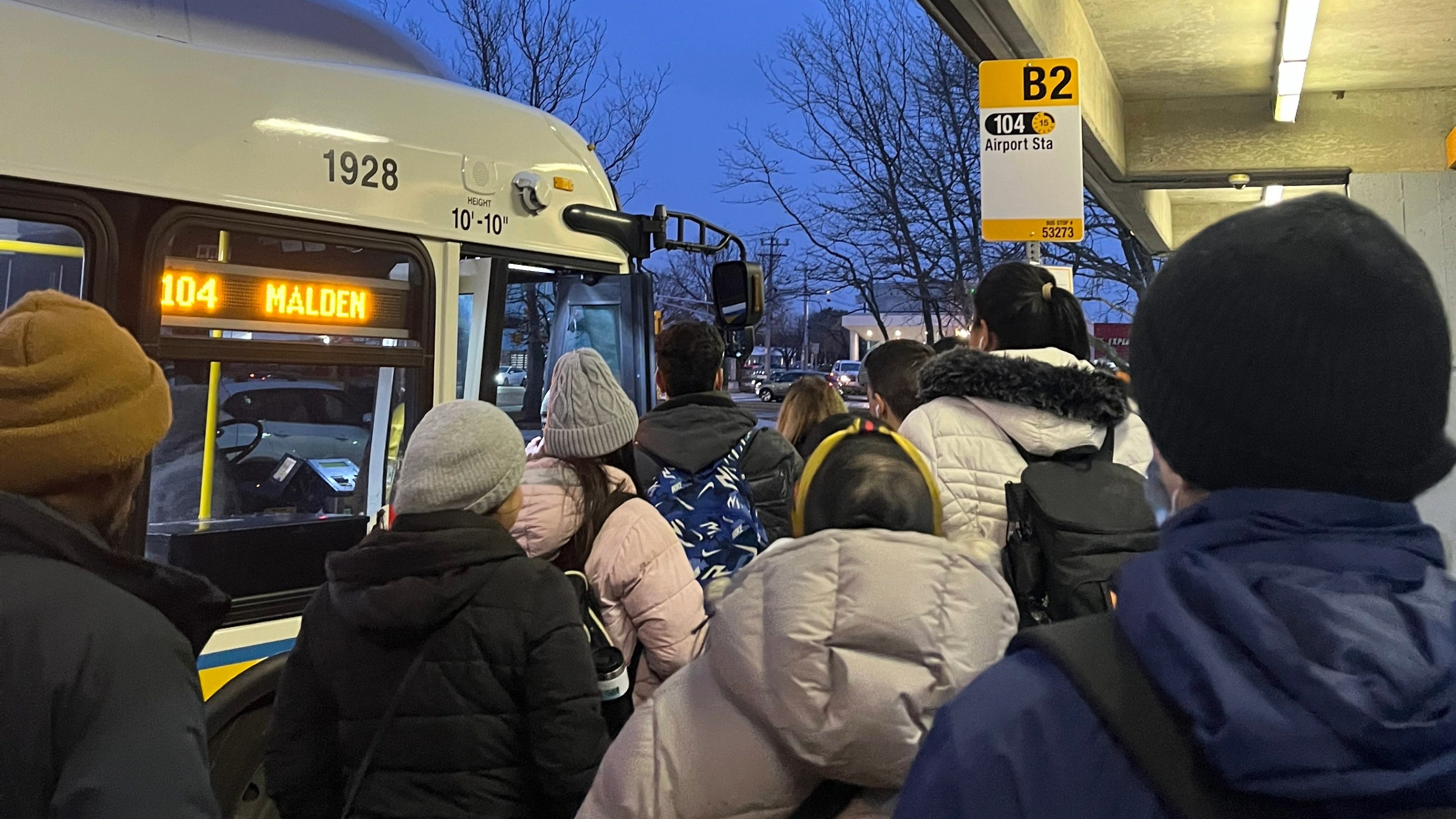 A crowd of people in winter coats waits to board an MBTA 104 bus.