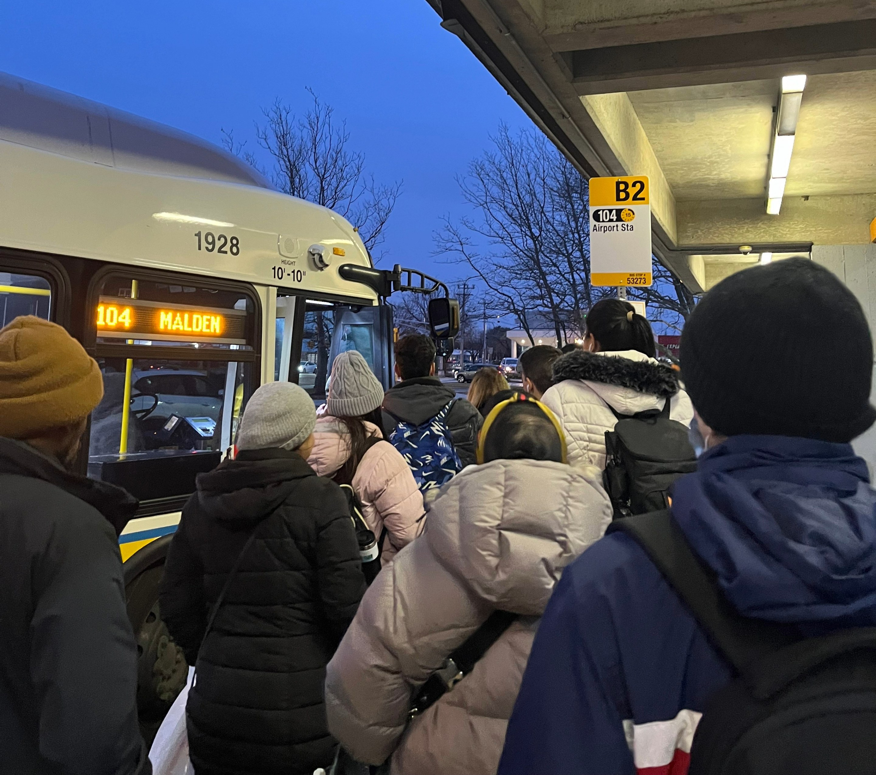 A crowd of people in winter coats waits to board an MBTA 104 bus.