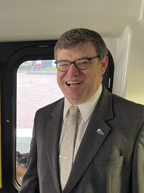 A middle-aged man in a suit stands in the operator cab of a light rail train.