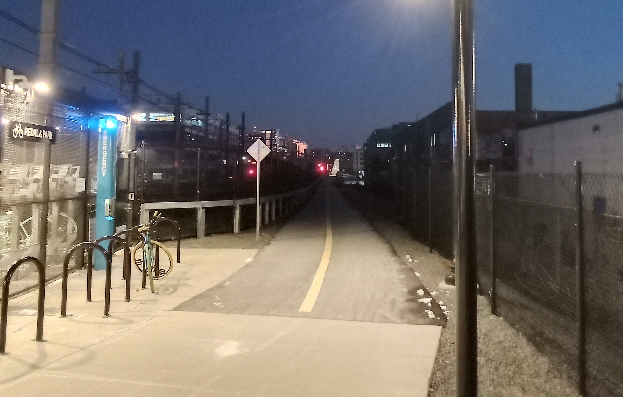 A brightly-lit bike path next to some bike racks extends into the distance on a long, unlit straightway in the dark (center) at dusk.