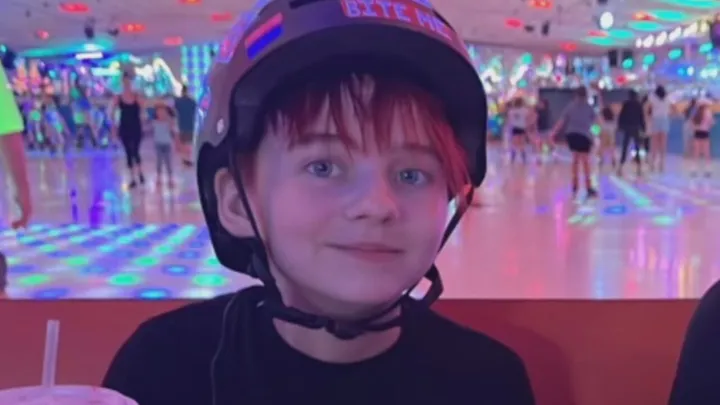 A blue-eyed kid with dyed pink bangs and a helmet grins in a roller rink.