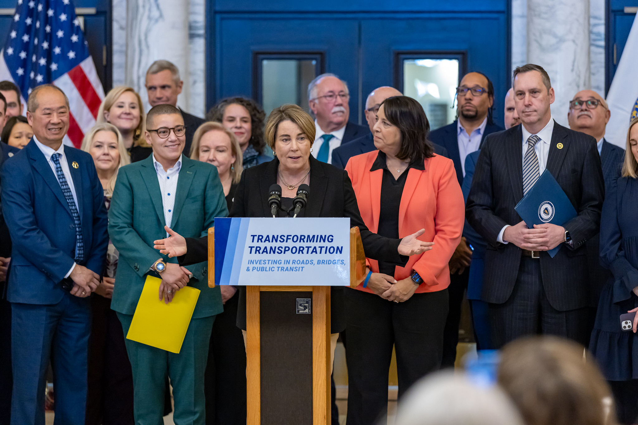 Governor Maura Healey stands in front of a crowd of government officials in suits behind a podium with a "Transforming Transportation" sign posted on its front.