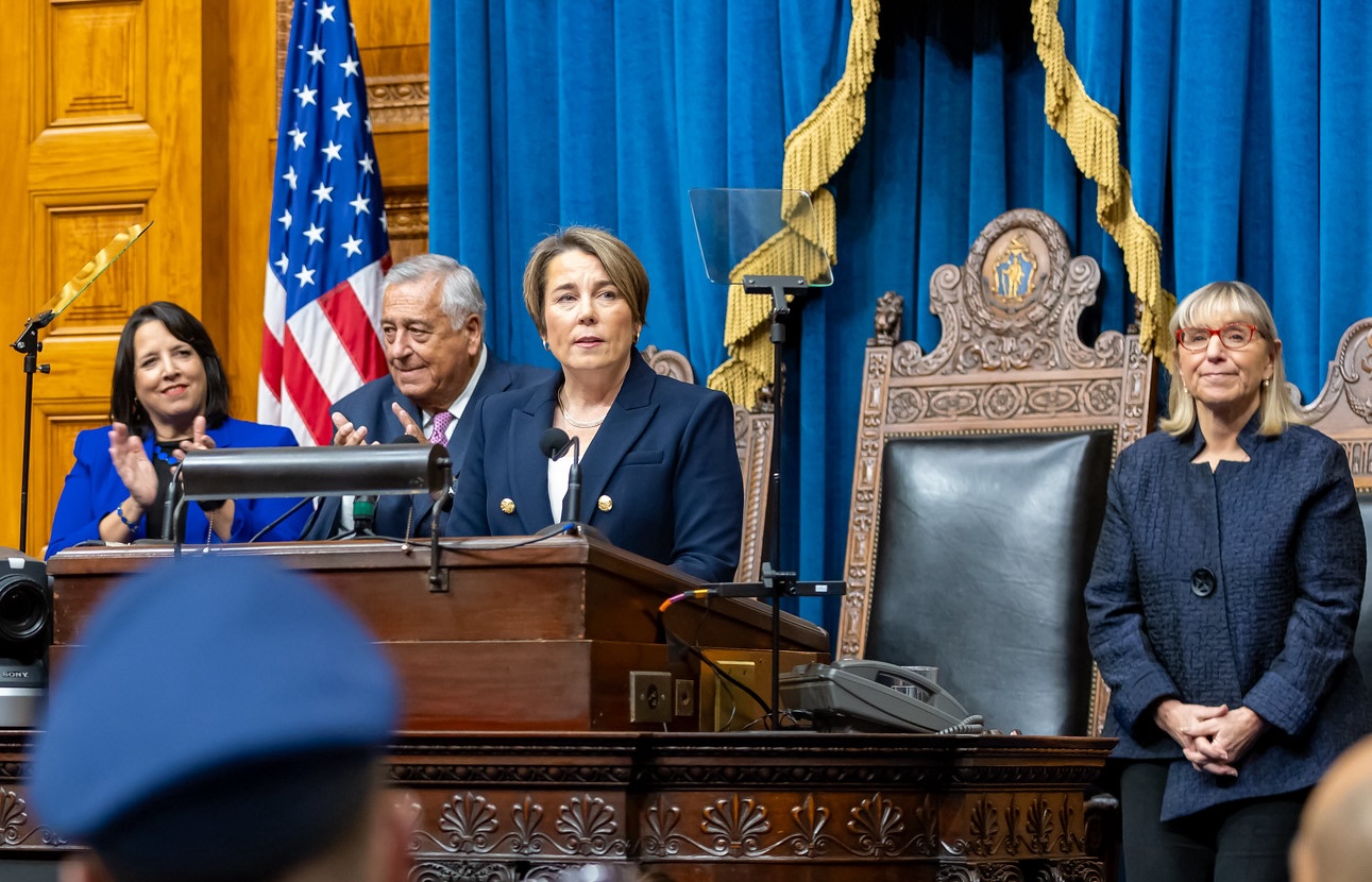 A woman with short brown hair stands at a podium in front of blue drapes, an American flag, and an elaborately carved wooden chair with the state seal of Massachusetts on its high back. Three other middle-aged people in suits are standing behind her.