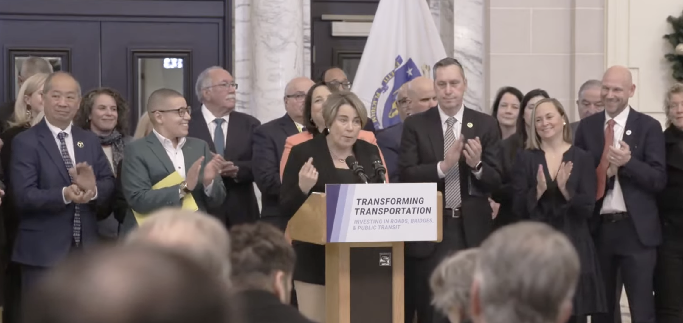 Governor Maura Healey stands in front of a crowd of applauding government officials behind a podium with a "Transforming Transportation" sign posted on its front.