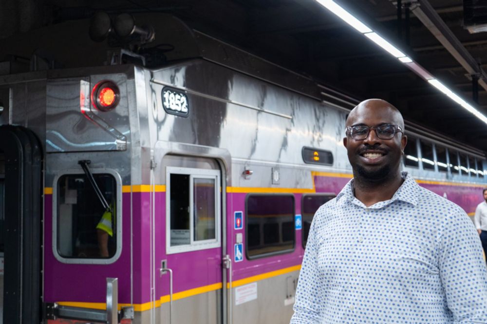 A black main in a collared shirt stands in front of a purple-and-silver MBTA commuter rail train.