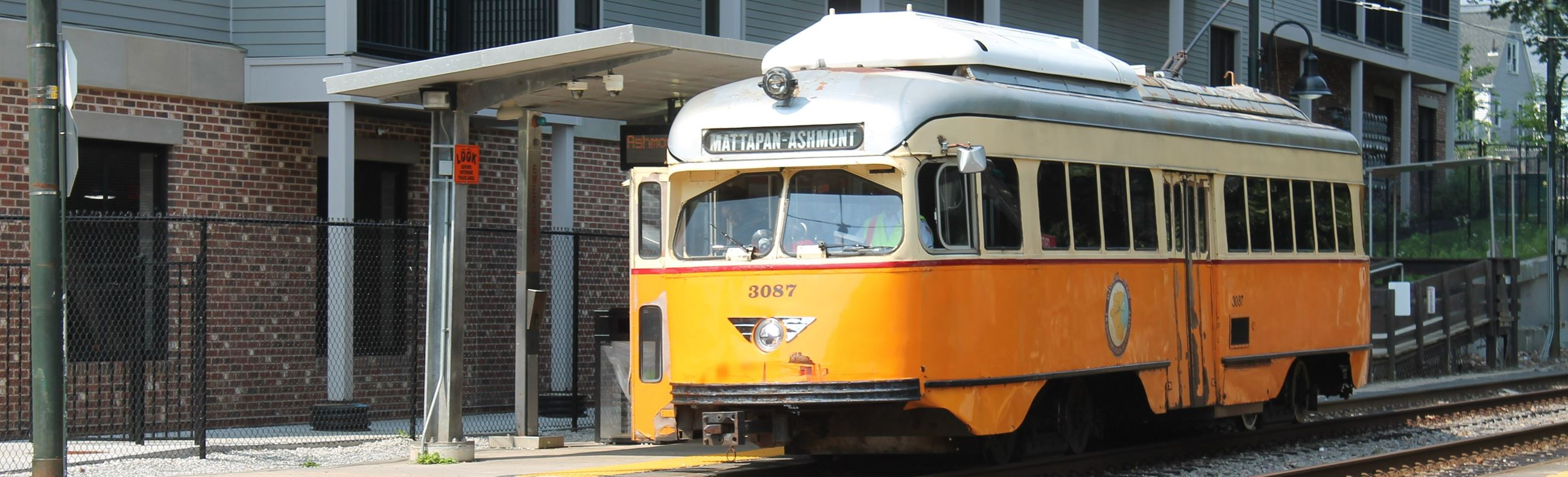 An orange vintage streetcar with the words "MATTAPAN-ASHMONT" on its destination sign stopped at a platform next to a building.