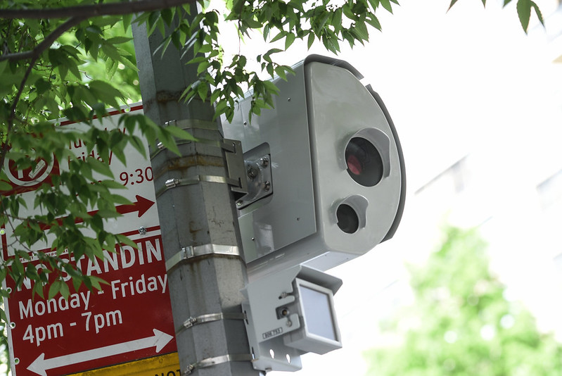 A camera inside a gray metal box mounted to a street light pole next to a "no standing" sign.