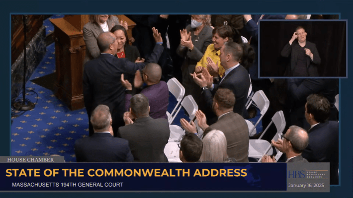 A group of people stands in a packed Massachusetts State House chamber. A man in a suit shakes hands with another individual while others clap and cheer. Chairs and wooden podiums are visible in the background. An inset of an ASL interpreter is on the screen.