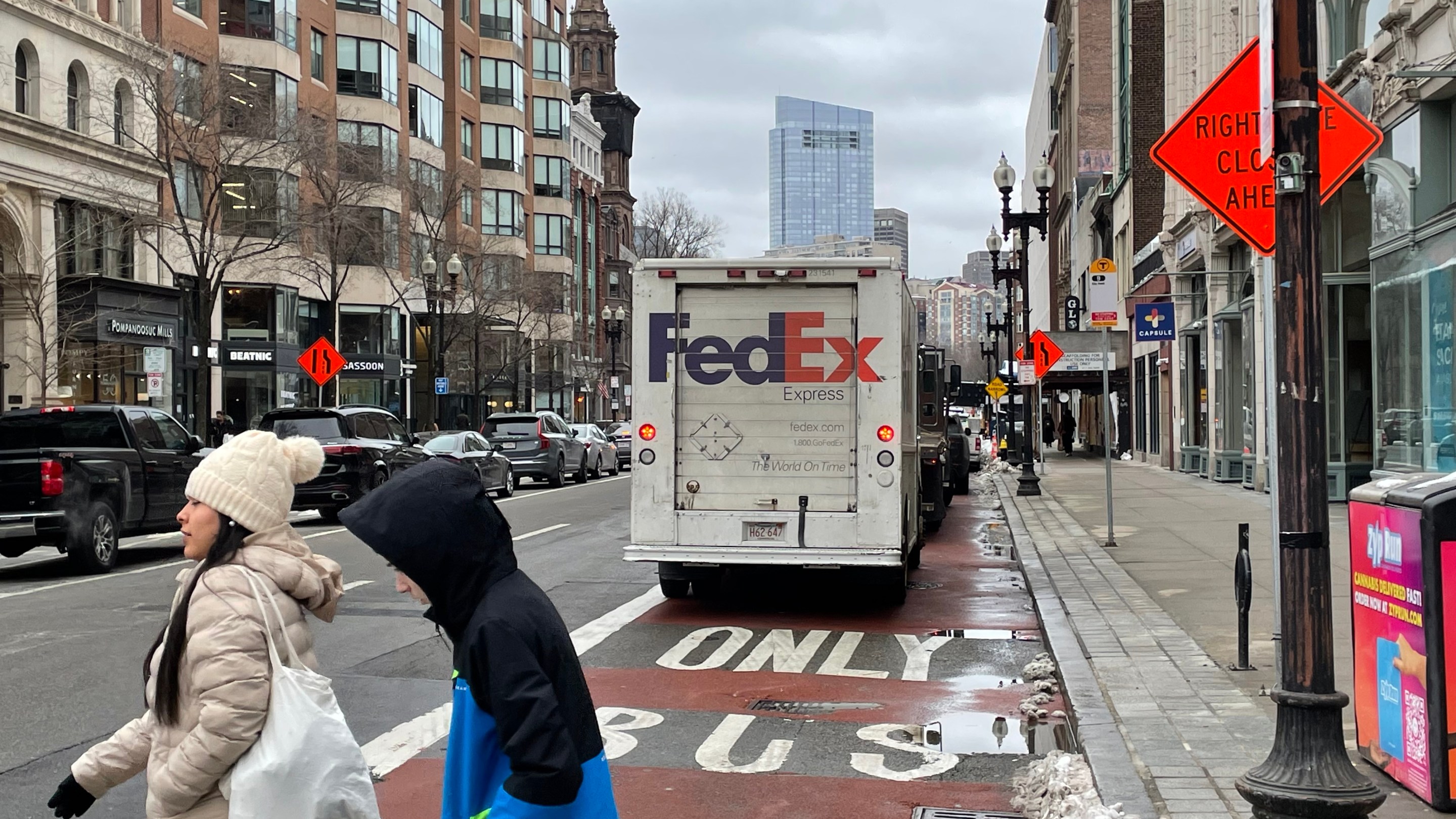 A FedEx truck and other illegally parked vehicles parked in the middle of a red bus lane marked with the words "ONLY BUS" on a city street lined with tall buildings. In the foreground, two people in winter coats cross the street.