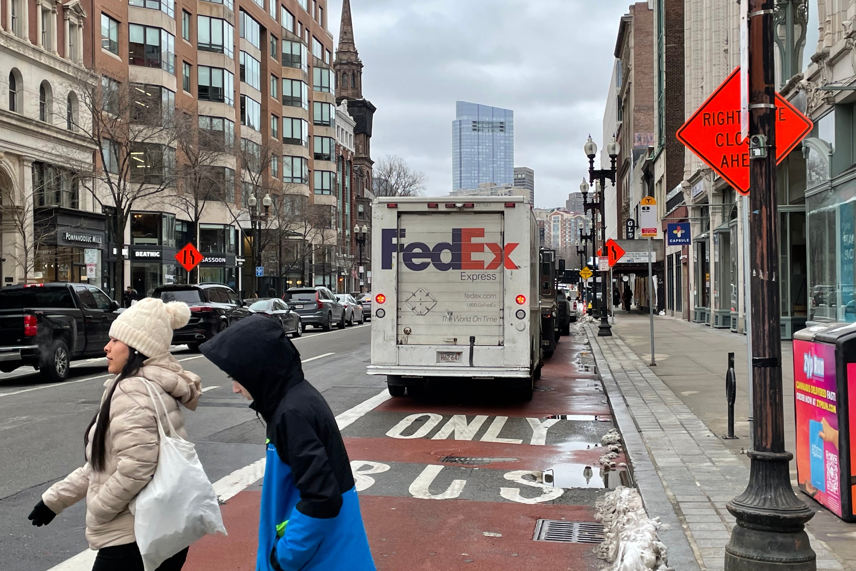 A FedEx truck and other illegally parked vehicles parked in the middle of a red bus lane marked with the words "ONLY BUS" on a city street lined with tall buildings. In the foreground, two people in winter coats cross the street.