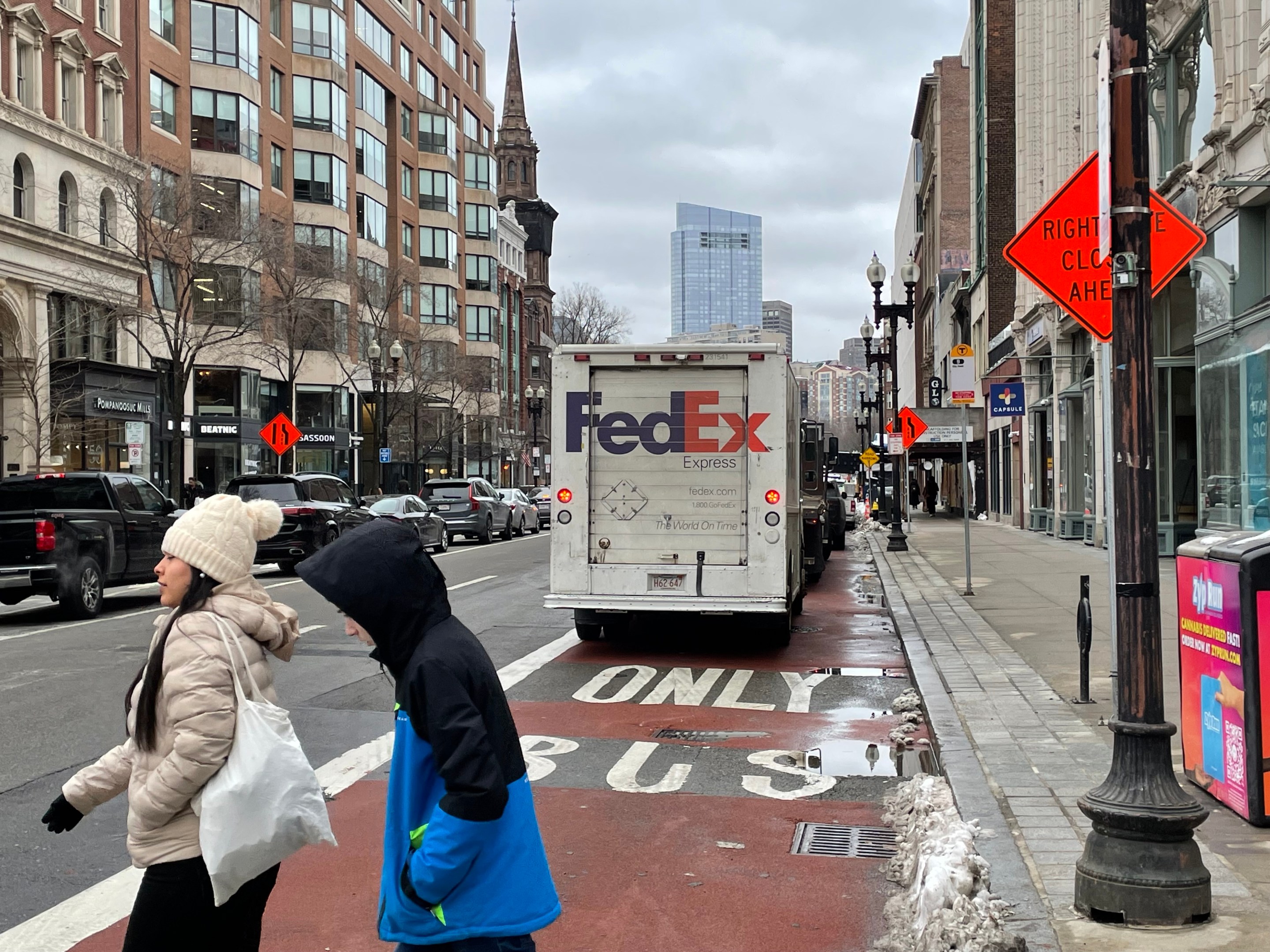 A FedEx truck and other illegally parked vehicles parked in the middle of a red bus lane marked with the words "ONLY BUS" on a city street lined with tall buildings. In the foreground, two people in winter coats cross the street.