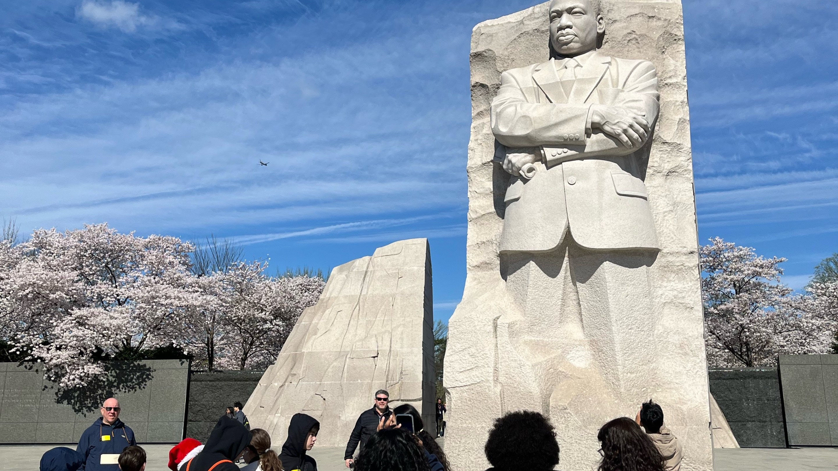 Visitors gather and look on at the Martin Luther King Jr. Memorial in Washington, D.C., admiring the impressive stone statue of the civil rights leader against a backdrop of cherry blossoms and a clear sky.