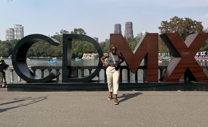 A smiling Black woman stands in front of a large sculpture of letters that spell "CDMX" next to a lake.