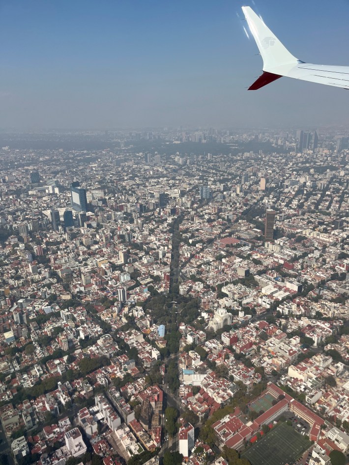 Aerial view of Mexico City with a plane wing visible in the top right corner. The cityscape is densely packed with buildings, and a prominent tree-lined avenue runs through the center of the image. The sky is clear with a slight haze in the distance.