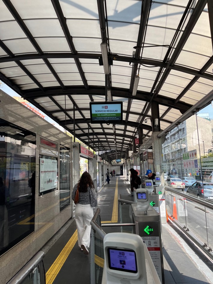 Faregates at the entrance to an enclosed center-of-the-street bus platform with glass walls and a curved roof. Several passengers are waiting beyond the fare gates