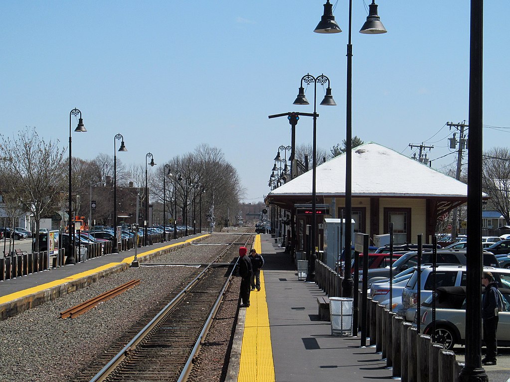 A view of a train platform next to a single railroad track in winter. A hip-roofed station building is visible next to the platform in the middle distance.