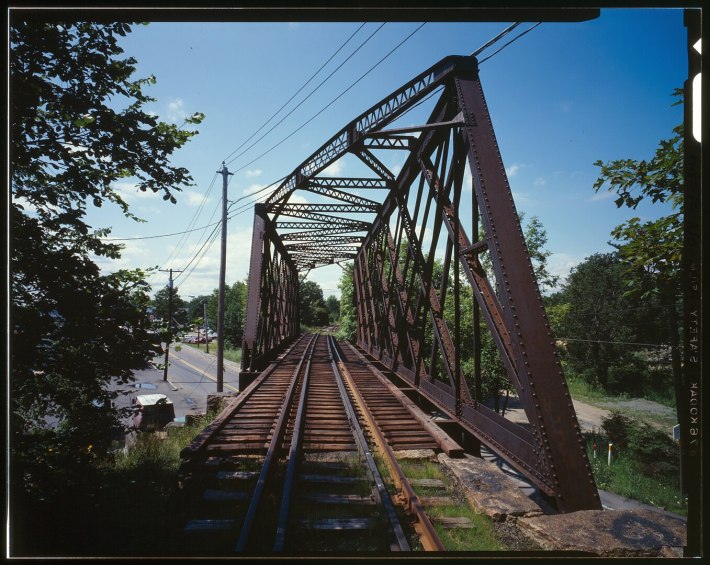 A view from the tracks of a steel truss railroad bridge over a city street in the summer time.