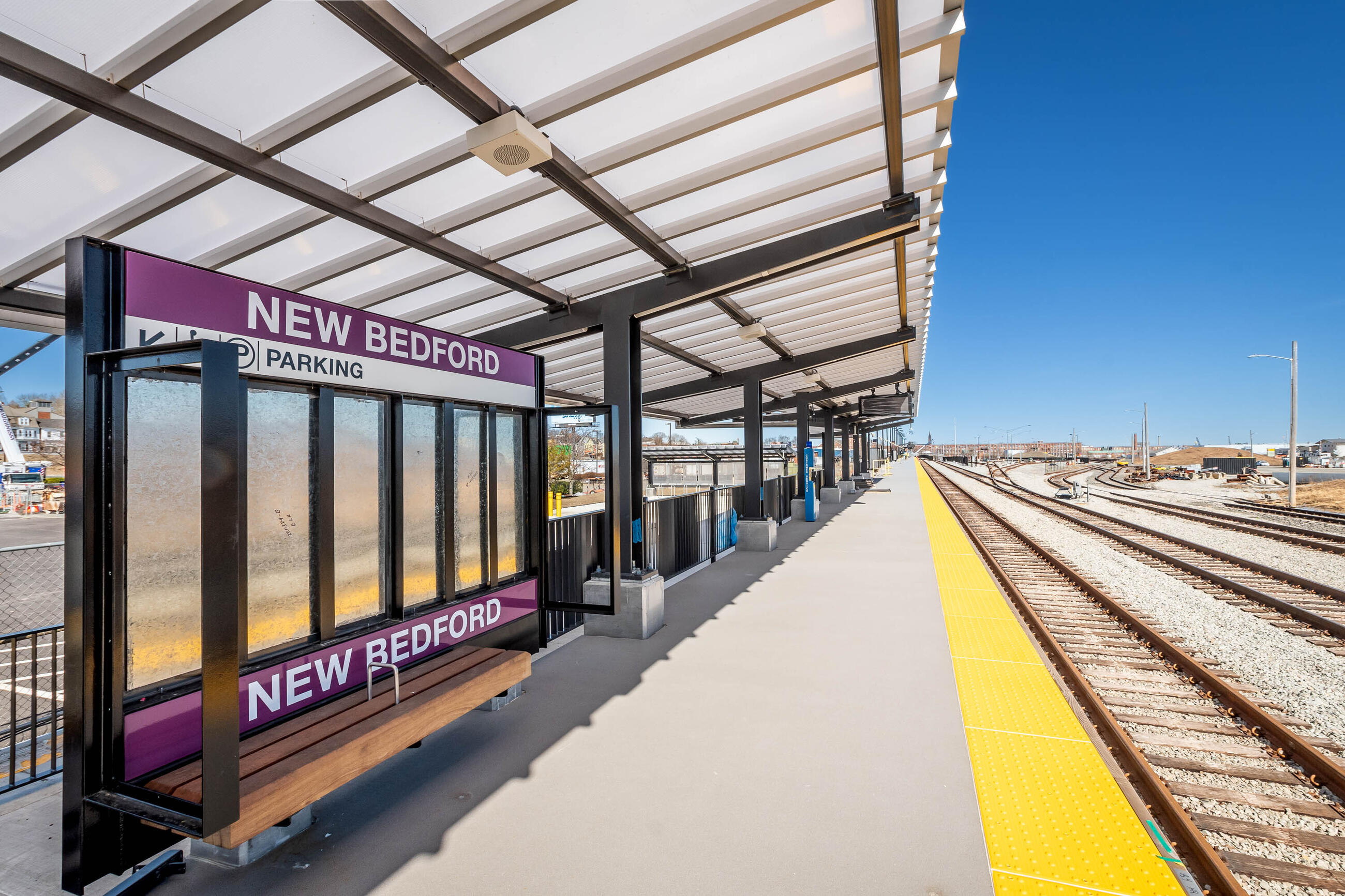An empty commuter rail platform with a shade canopy overhead. A sign under the canopy reads "NEW BEDFORD" in white text on a purple background. To the right are four sets of railroad tracks leading into the distance.