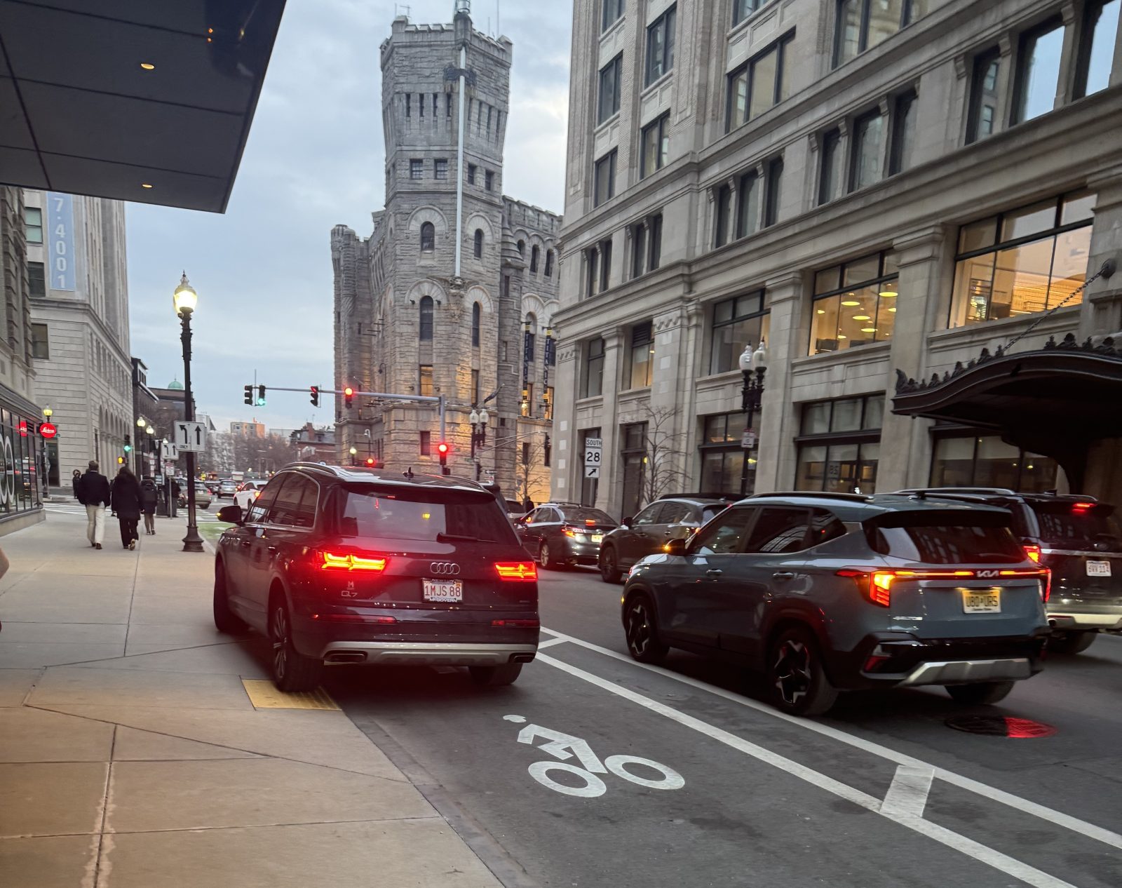 An illegally parked minivan blocks both the bike lane and and a sidewalk curb ramp on a multi-lane city street lined with historic masonry buildings.