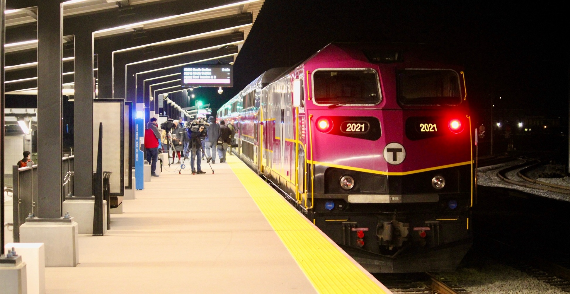 A crowd of passengers on a lit train platform under a dark sky, with a purple MBTA commuter rail train at right.