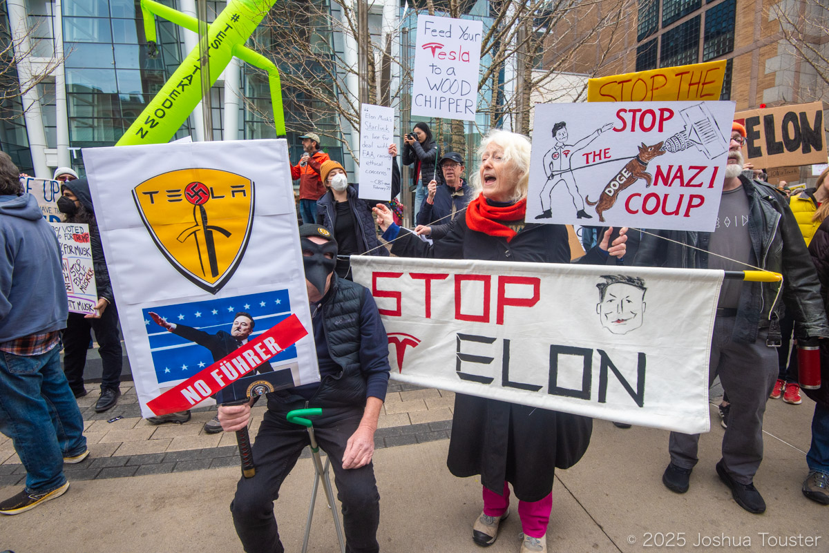 A crowd of protesters in winter clothing hold signs that say "Stop Elon" and "Tesla – No Fuhrer" and "Stop the Nazi coup" and "feed your Tesla to a wood chipper"