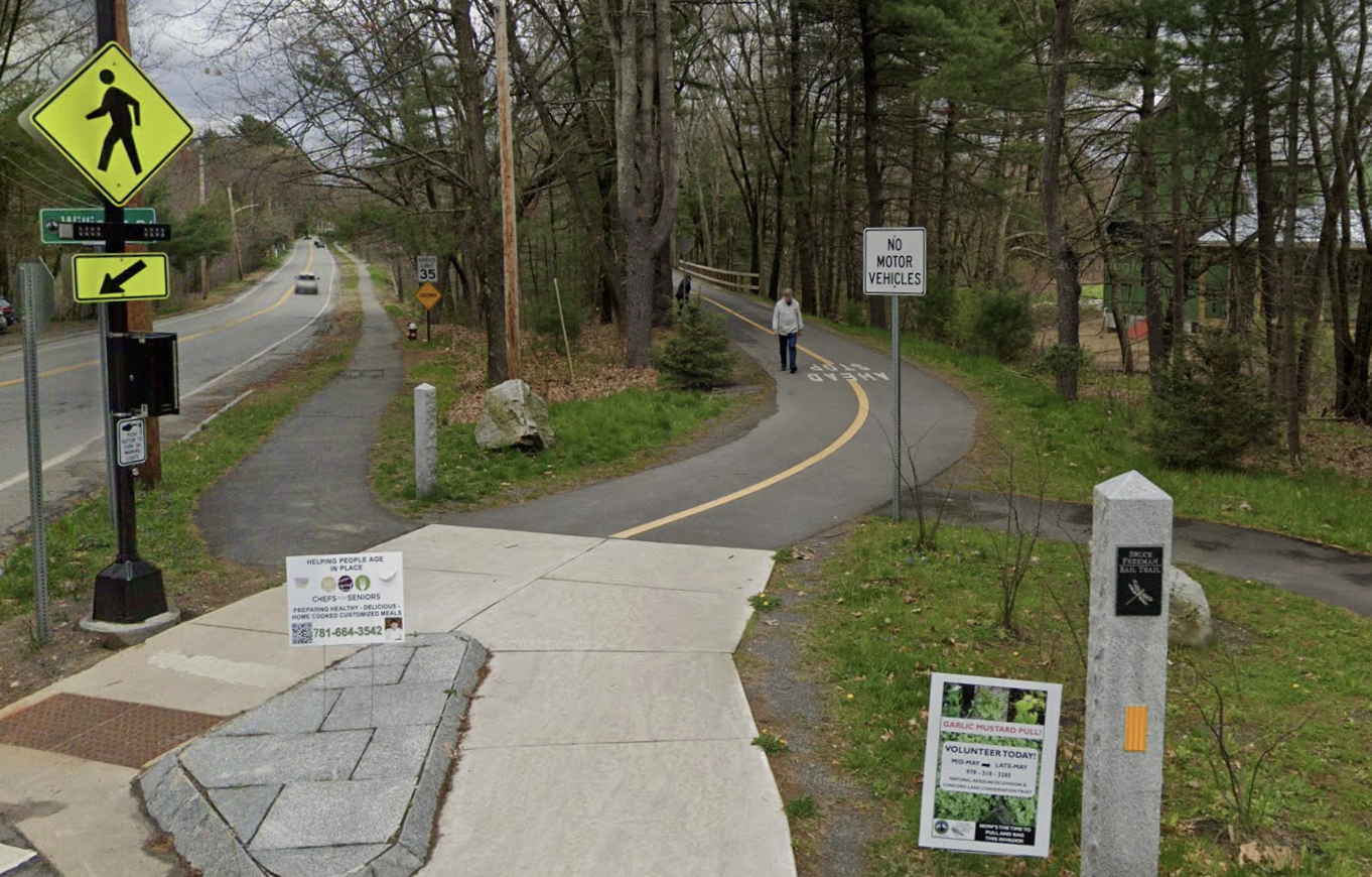 A bike trail entrance in the woods next to a rural two-lane roadway. A yellow pedestrian crossing sign is visible at upper left.