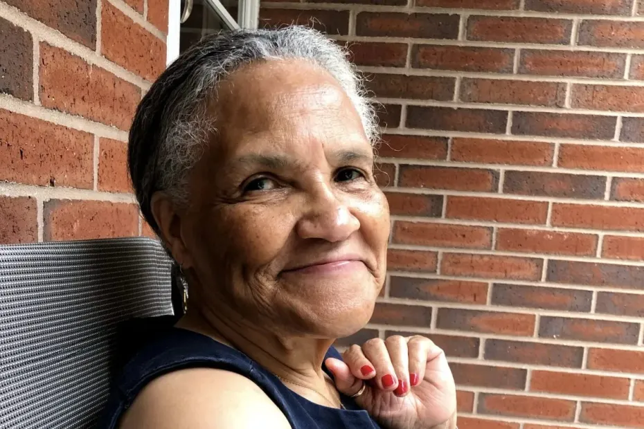 An elderly, grinning brown-skinned woman sitting next to a brick wall