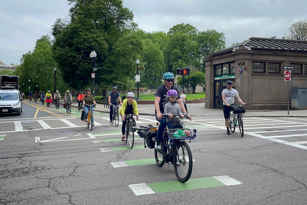 A long line of people on bikes rides along a bike lane through an intersection. In the background is a large park with a subway station entrance on the corner. A green sign on the subway station reads "BOYLSTON". In the front of the line is a man in a teal bike helmet carrying a young child in a purple helmet on a kids seat in front of him.